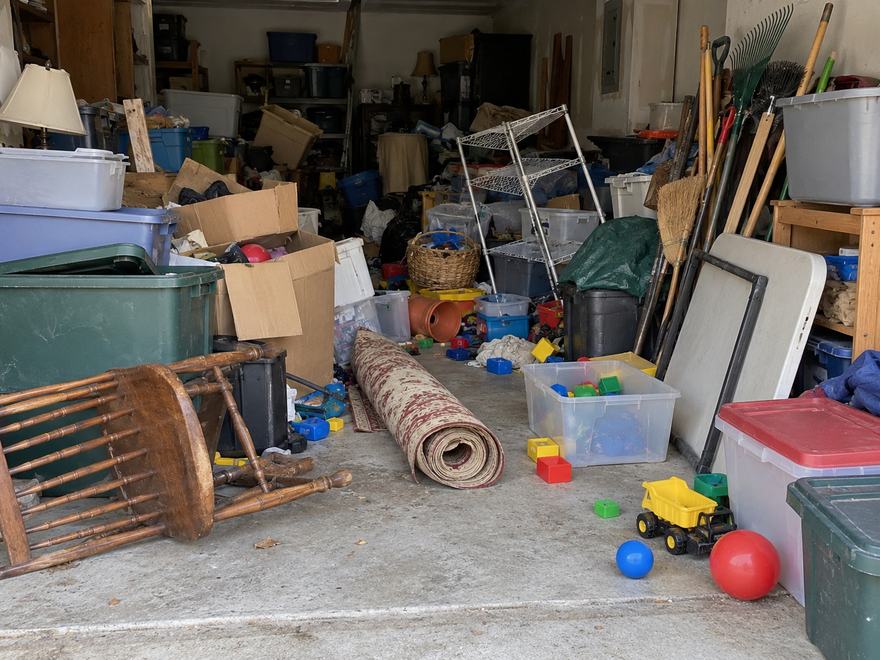 Cluttered garage filled with storage bins, cardboard boxes, toys, a rolled rug and garden tools before a cleanout